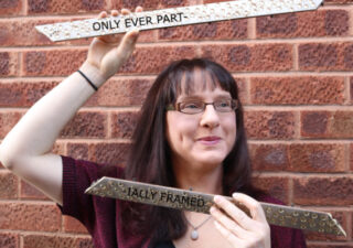 Photo of Sarah Leavesley standing in front of a brick wall. She is holding two bits of a picture frame. Oh! She's framed! But the bits of frame have words on ... 'Only ever part-' on the top one and '-ially framed' on the bottom one. Oh, I get it now. Clever.