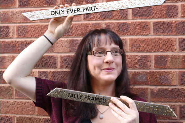 Photo of Sarah Leavesley standing in front of a brick wall. She is holding two bits of a picture frame. Oh! She's framed! But the bits of frame have words on ... 'Only ever part-' on the top one and '-ially framed' on the bottom one. Oh, I get it now. Clever.