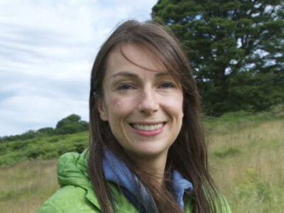 Photo of Zoë Walkington. She has long dark hair and is smiling. She is standing in a field with trees behind her.