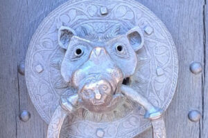 Close up of a doorknocker on a large old wooden door. The knocker is metal and shows a pig or boar with a ring in its mouth.