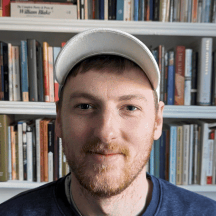 Dane Holt in front of a bookshelf, he has a beard and a baseball cap
