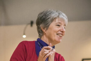 Photograph of Helen Evans. She has short grey hair and is clasping her hands and leaning to her left.