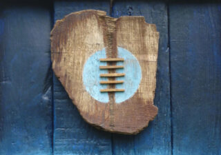 A wooden sculpture hung on an old blue wooden plank door. The brown, roughly heart shaped and has a light blue circle in the centre crossed with what look like laces.
