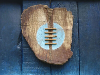 A wooden sculpture hung on an old blue wooden plank door. The brown, roughly heart shaped and has a light blue circle in the centre crossed with what look like laces.