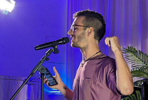 A young man with a small beard is reading poetry into a microphone, reading from a mobile phone and waving a clenched fist.