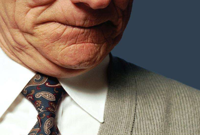 A photograph of a man wearing a spotless white shirt, a paisley tie and a beige cardigan. Only the lower half of the face is visible,