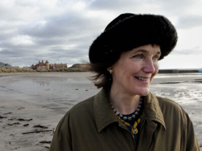 Photograph of Katrina walking on a beach wearing a round black hat. (photo Joe Grabham).