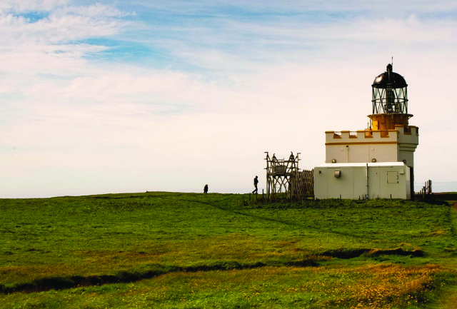 A landscape photograph showing grass, a cloudy sky and a small crenelated building with a tower that looks like a lighthouse, but is presumably a camera obscura