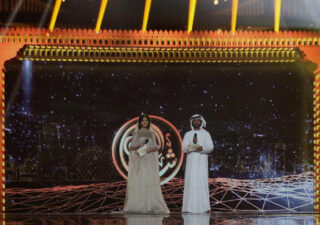 A fancy proscenium arch on a stage in gold and red. Underneath on the stage are a man and a woman in Arabic dress.