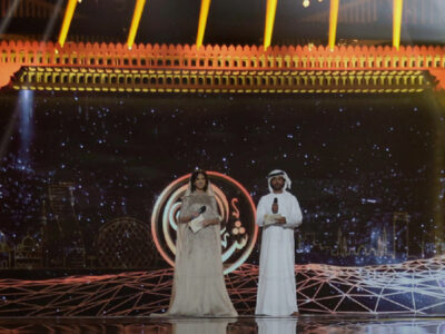 A fancy proscenium arch on a stage in gold and red. Underneath on the stage are a man and a woman in Arabic dress.