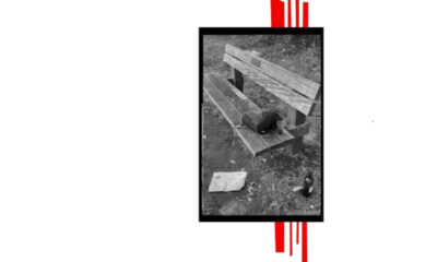 black and white photograph of a park bench. There is a small backpack on one end. A glass and a bottle are nearby.